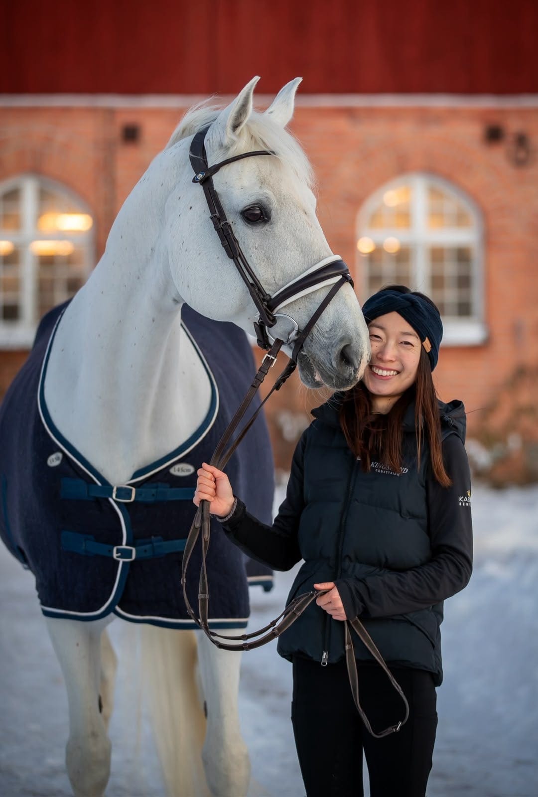 Yuka smiling next to a grey horse in a winter blanket